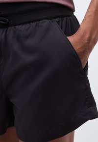 Κοντινό πλάνο ενός ατόμου που φοράει μαύρο σορτς με ελαστική μέση και έχει το χέρι του στην δεξιά τσέπη.