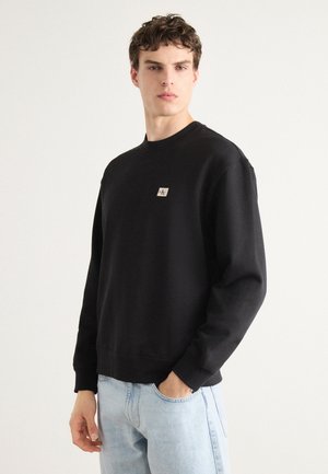 Black sweatshirt made of soft fabric, featuring a round neckline, long sleeves, and a small logo patch on the chest. Paired with light blue jeans.