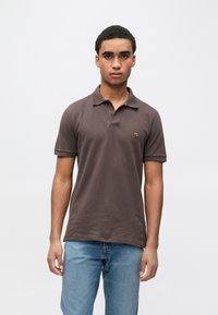 Homem jovem em pé de frente, vestindo uma camisa pólo castanha de mangas curtas e jeans azul claro, contra um fundo branco liso.