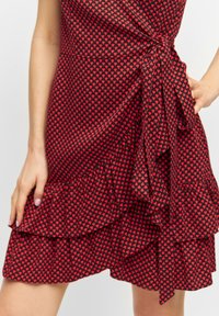 Robe rouge avec un petit motif de cœurs noirs, ornée de volants au bas et d'un détail de taille nouée. Fabriquée en tissu léger.