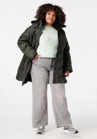 Pufferjack in donkergroen met een glanzende textuur, gecombineerd met een licht muntgroene trui met pijlpatronen en relaxed-fit grijze jeans.