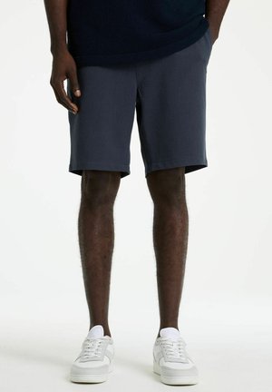 Männliches Model trägt marineblaue knielange Shorts und weiße Sneaker, steht mit den Händen in den Taschen vor schlichtem Hintergrund.