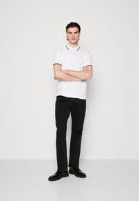 Weißes Poloshirt mit schwarzem Rand, schwarze Jeans und schwarze Schuhe. Das Poloshirt hat einen Kragen und zwei Knöpfe. Das Modell steht mit verschränkten Armen.