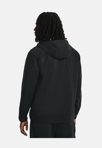 Veste à capuche noire avec une texture lisse et une coupe décontractée. Elle présente un col rond, des manches longues et des détails de couture simples.