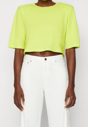 Osoba ubrana w limonkowo zielony krótki t-shirt w połączeniu z białymi, wysokimi dżinsami, ręce swobodnie ułożone wzdłuż ciała na jednolitym tle.