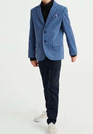 Blauer Cordblazer mit zwei Knöpfen, Reverskragen und einem Einstecktuch, kombiniert mit dunklen Hosen und hellen Sneakers. Strukturiertes Material.