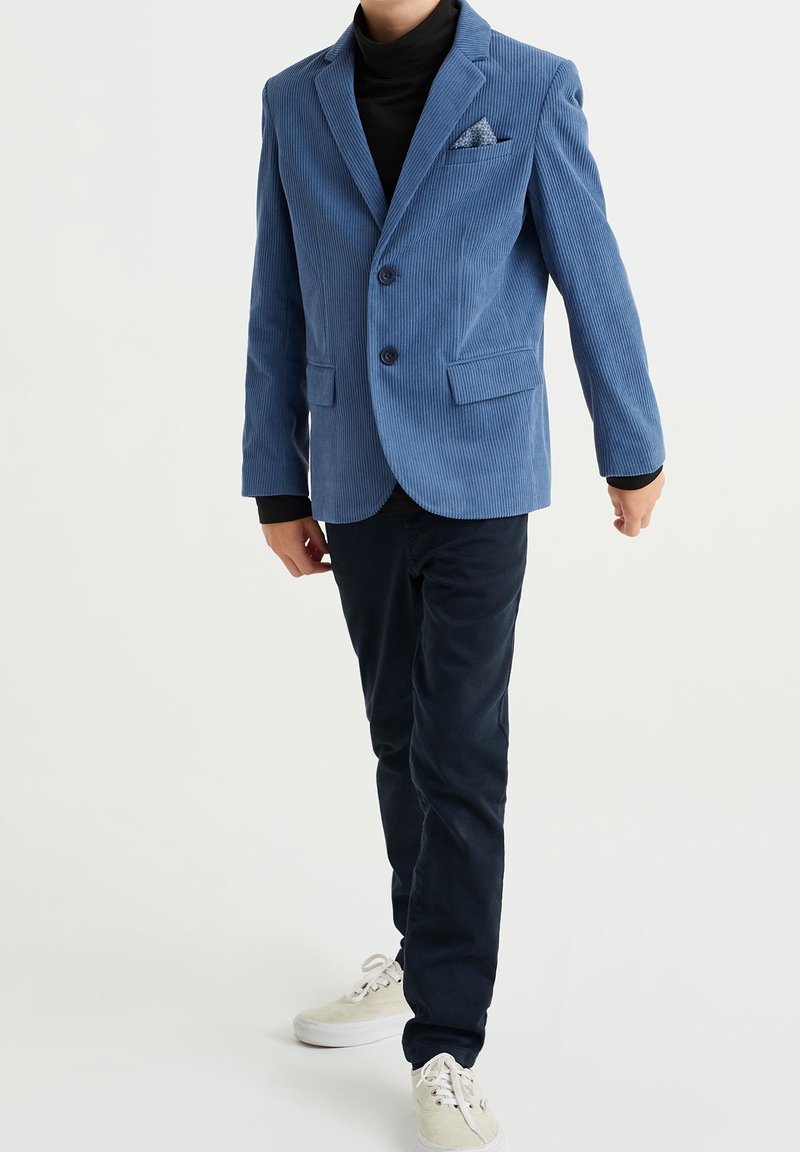 Modrý kordový blazer se dvěma knoflíky, klopami a kapesníčkem, spárovaný s tmavými kalhotami a světlými teniskami. Texturovaná látka.