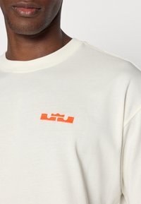 Krem-sivi dukserica s okruglim izrezom, s malim narančastim logotipom na prsima. Napravljen od mekog, teksturiranog materijala.