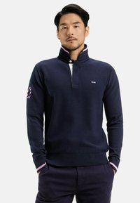 Pull en maille bleu marine avec un col haut, présentant un col intérieur blanc, des accents de rayures roses et un petit logo sur la poitrine.