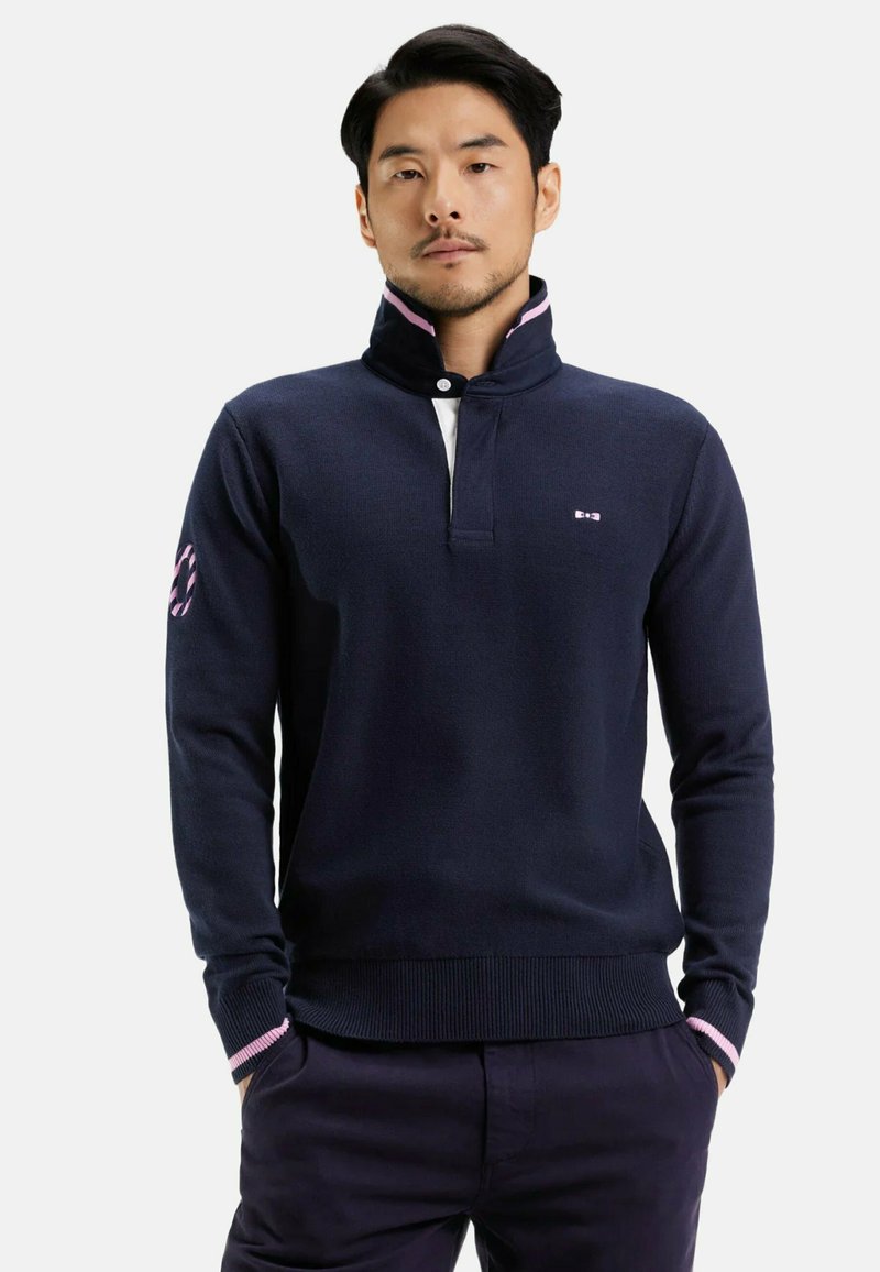 Pull en maille bleu marine avec un col haut, présentant un col intérieur blanc, des accents de rayures roses et un petit logo sur la poitrine.