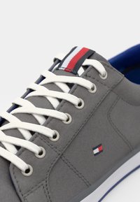 Zapatilla deportiva de lona gris con cordones blancos, interior azul y un logotipo con detalles en rojo, blanco y azul en la lengüeta.