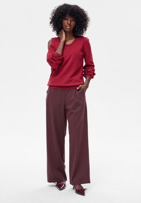 Czerwony sweter z dzianiny z szerokimi mankietami i zaokrąglonym dekoltem, zestawiony z luźnymi bordowymi spodniami i butami na obcasie z noskiem w szpic. Gładkie faktury na całej powierzchni.