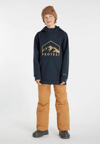 Felpa con cappuccio impermeabile blu navy con logo di una montagna dorata, abbinata a pantaloni da neve marroni e stivali da neve neri. Berretto lavorato a maglia marrone in testa.