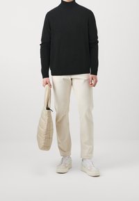 Person in einem schwarzen Rollkragenpullover, hellbeigen Hosen und weißen Sneakern, die eine beige, gesteppte Handtasche vor einem schlichten hellen Hintergrund hält.