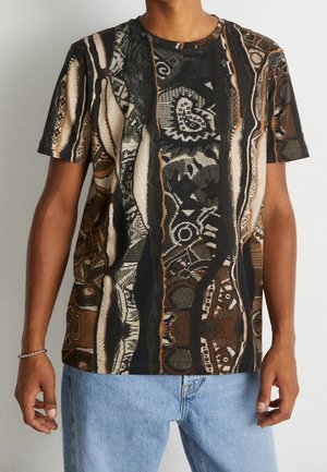 Persoon in een shirt met korte mouwen in bruin, zwart en beige, met abstracte ontwerpen, gecombineerd met lichtblauwe jeans.