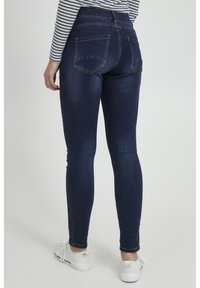 Jeans ajustados de mezclilla azul oscuro, con cintura alta, bolsillos traseros y costuras en contraste. Combinados con zapatillas blancas y una camiseta a rayas.