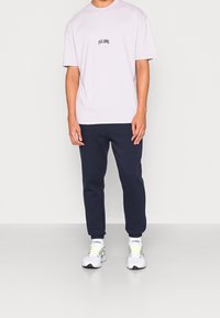 Světlé fialové tričko s krátkým rukávem a černým textem, spárované s tmavě modrými tepláky a bílými teniskami s žlutými akcenty.