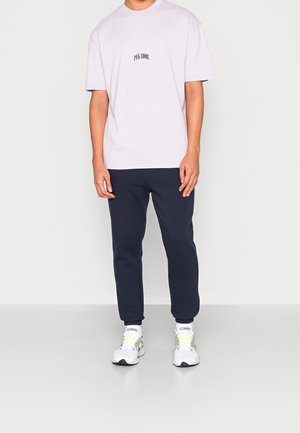 T-shirt à manches courtes violet clair avec du texte noir, associé à un pantalon de survêtement bleu marine et des baskets blanches avec des accents jaunes.