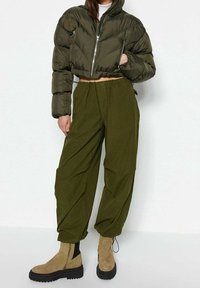 Nafukovaná olivově zelená krátká bunda, volné olivově zelené kalhoty a béžové kotníkové boty s černými podrážkami a bočními zipy na neutrálním pozadí.