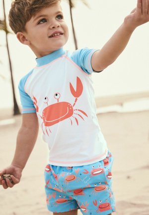 Il set di costumi da bagno per ragazzi include una maglietta bianca a maniche corte con accenti blu e una grafica di granchio, abbinata a pantaloni corti blu con un motivo di granchi.