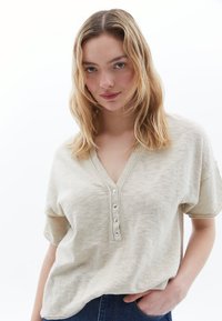 Beige Kurzarmoberteil mit V-Ausschnitt, Knopfleiste mit fünf Knöpfen und weicher Textur; getragen mit dunklen Jeans.