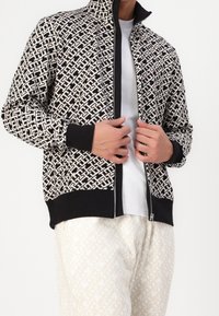 Persona que lleva una chaqueta con patrón en blanco y negro, con cremallera, sobre una camisa blanca, combinada con pantalones claros con patrones, ajustándose la chaqueta con las manos.