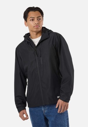 Jonge man draagt een zwart windjack met capuchon over een wit shirt en blauwe spijkerbroek, kijkt iets naar de zijkant.