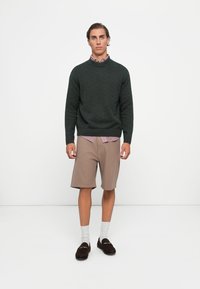 Jaunas vyras stovi tiesiai, vilkėdamas tamsiai žalią tekstūruotą megztinį, dryžuotą marškinėlį, smėlio spalvos šortus, baltas kojines ir tamsias loafer avalynes ant vienspalvės baltos fono.