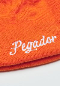 Lys orange strikket hue med hvid broderet tekst "Pegador" langs kanten. Teksten har en kursiv stil mod den levende baggrund.