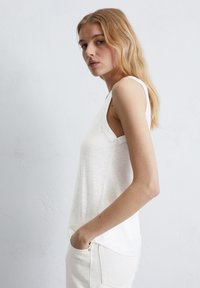 Haut sans manches blanc en tissu doux et léger avec une coupe décontractée. Présente un col rond et des détails sur les coutures latérales. À associer avec un pantalon de couleur claire.