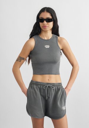 Γυναίκα που φοράει γκρι σκουρόχρωμο αμάνικο κοντό τοπ και αντίστοιχα σορτς με κορδόνι στη μέση, μαύρα ορθογώνια γυαλιά ηλίου, στέκεται με τα χέρια στις τσέπες.