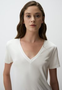 Biała koszulka z dekoltem w serek, wykonana z miękkiego, gładkiego materiału. Krótkie rękawy i czyste linie. Dekolt ma subtelną lamówkę, co tworzy minimalistyczny wygląd.