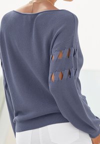 Blauer Strickpullover mit offenem Gitterdesign an den Ärmeln, gerippter Textur und lässiger Passform. Getragen mit weißen Hosen.