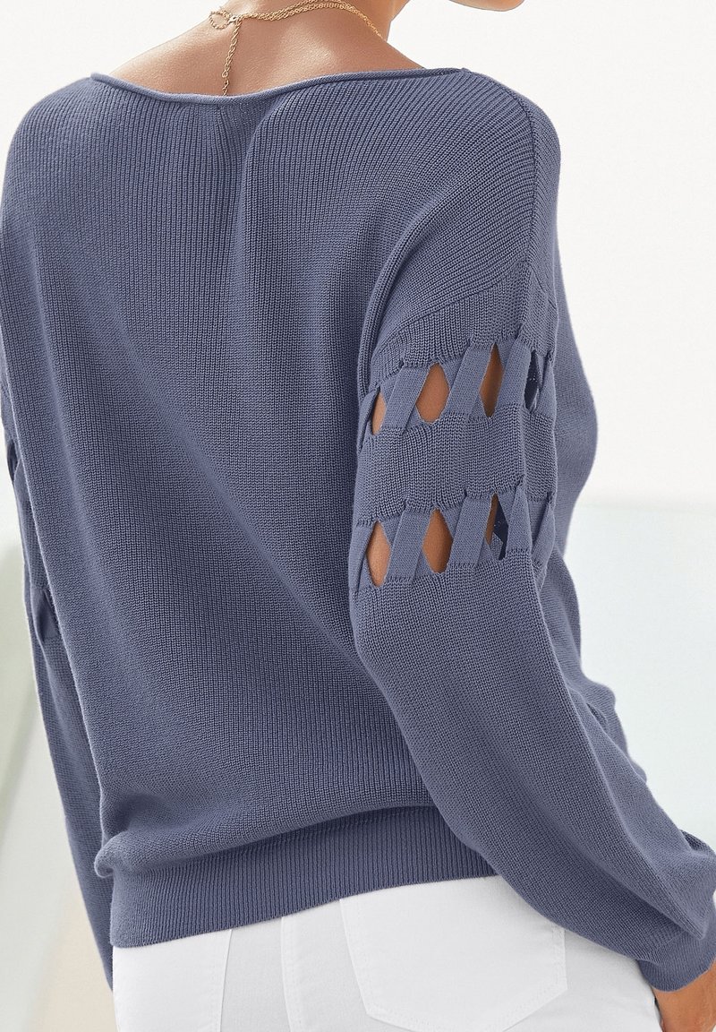 Blauer Strickpullover mit offenem Gitterdesign an den Ärmeln, gerippter Textur und lässiger Passform. Getragen mit weißen Hosen.