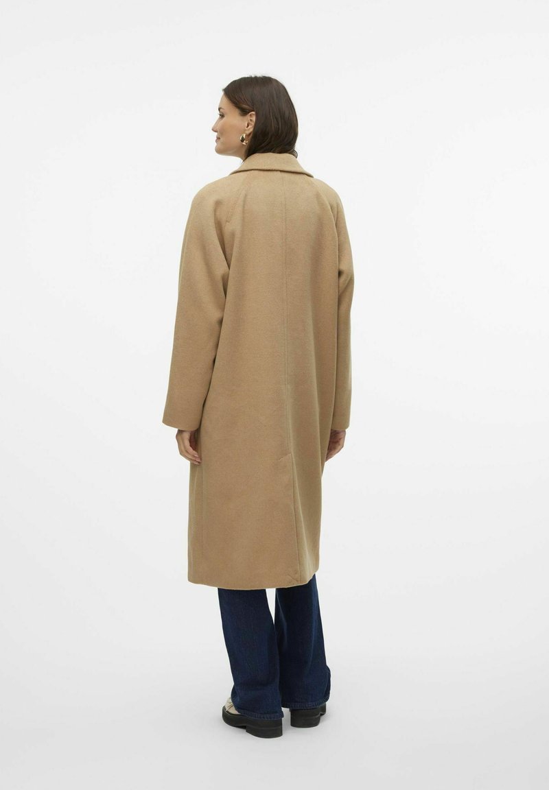 Manteau en laine brun clair, mi-long, avec un col et des détails de couture au dos, associé à un pantalon en jean foncé évasé et des bottes.