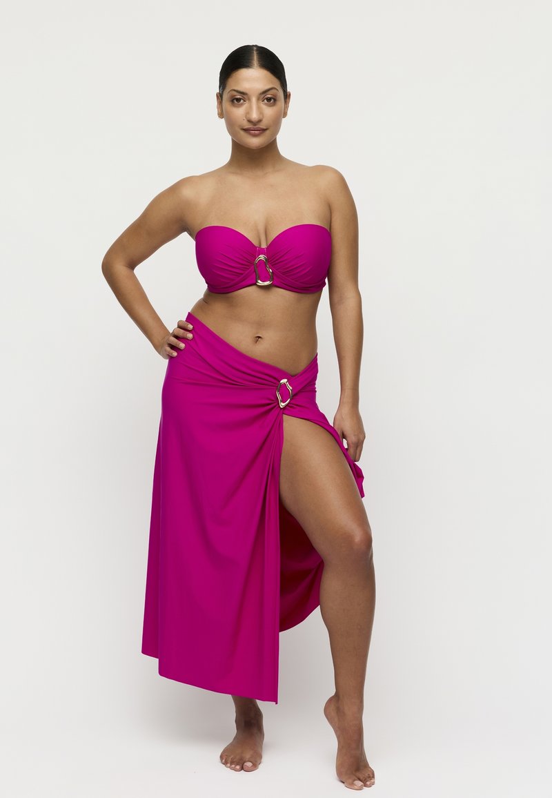 Frau mit dunklen Haaren, die ein trägerloses magentafarbenes Bikinitop und einen passenden Rock mit Seitenschlitz und dekorativen Ringen trägt, steht barfuß.