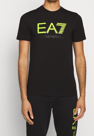 Homme portant un t-shirt noir EA7 Emporio Armani avec un logo jaune fluo et un pantalon noir assorti avec un motif jaune fluo sur la cuisse.
