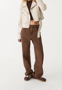 Veste de motard en simili cuir beige clair, cropped, avec des accessoires en argent, associée à un pantalon large taille haute marron et des baskets marron.