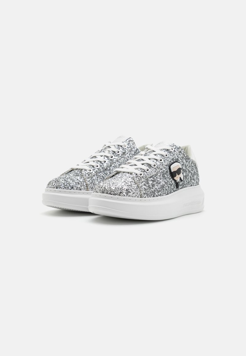 Zapatillas plateadas brillantes con suela blanca, que cuentan con cordones blancos y un acento gráfico negro en el costado. Parte superior texturizada, diseño moderno.