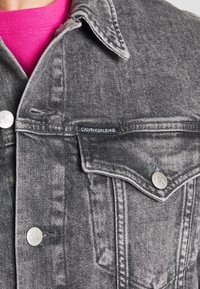 Szara kurtka dżinsowa z srebrnymi zatrzaskami, metką z napisem "CALVIN KLEIN JEANS" oraz teksturowanym wykończeniem. Nosi się ją na różową koszulę.