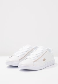 White lace-up sneakers with a smooth leather finish, accented by a beige stripe and a gold logo on the side. Rubber sole.