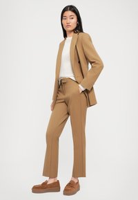 Femme se tient les mains dans les poches, portant un blazer et un pantalon couleur camel, un pull blanc et des mocassins compensés marron assortis.