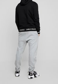 Pantaloni grigi in felpa con vestibilità comoda, dotati di un cinturino nero con il logo "CARLO COLUCCI". Abbinati a una felpa con cappuccio nera e sneakers nere.