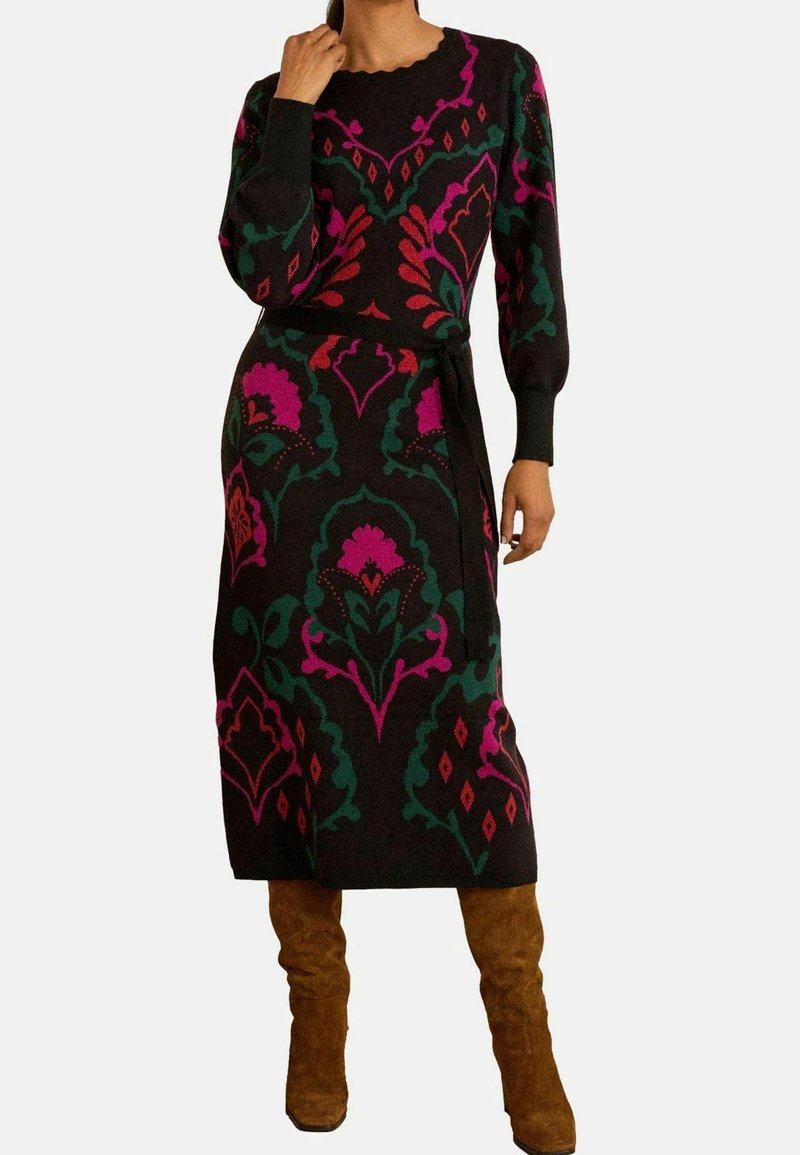 Femme portant une robe midi noire ornée de motifs floraux audacieux rose et vert, associée à des bottes montantes en daim marron.