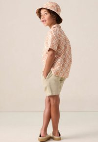 Kortarmige, lichtoranje gemusterde shirt gecombineerd met beige shorts. Een bijpassende gemusterde bucket hat maakt de outfit compleet. Schoeisel is een casual instapper.
