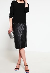 Pull noir à manches longues avec des accents décoratifs en forme d'étoile au bas, associé à une jupe en cuir noir ajustée. Pochette argentée et sandales noires.