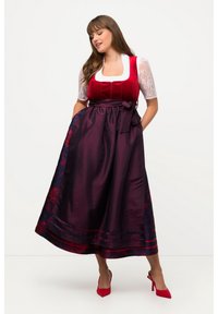 Frau in einem traditionellen roten und lila Dirndlkleid mit Spitzenärmeln und roten High Heels, die mit den Händen in den Taschen steht.