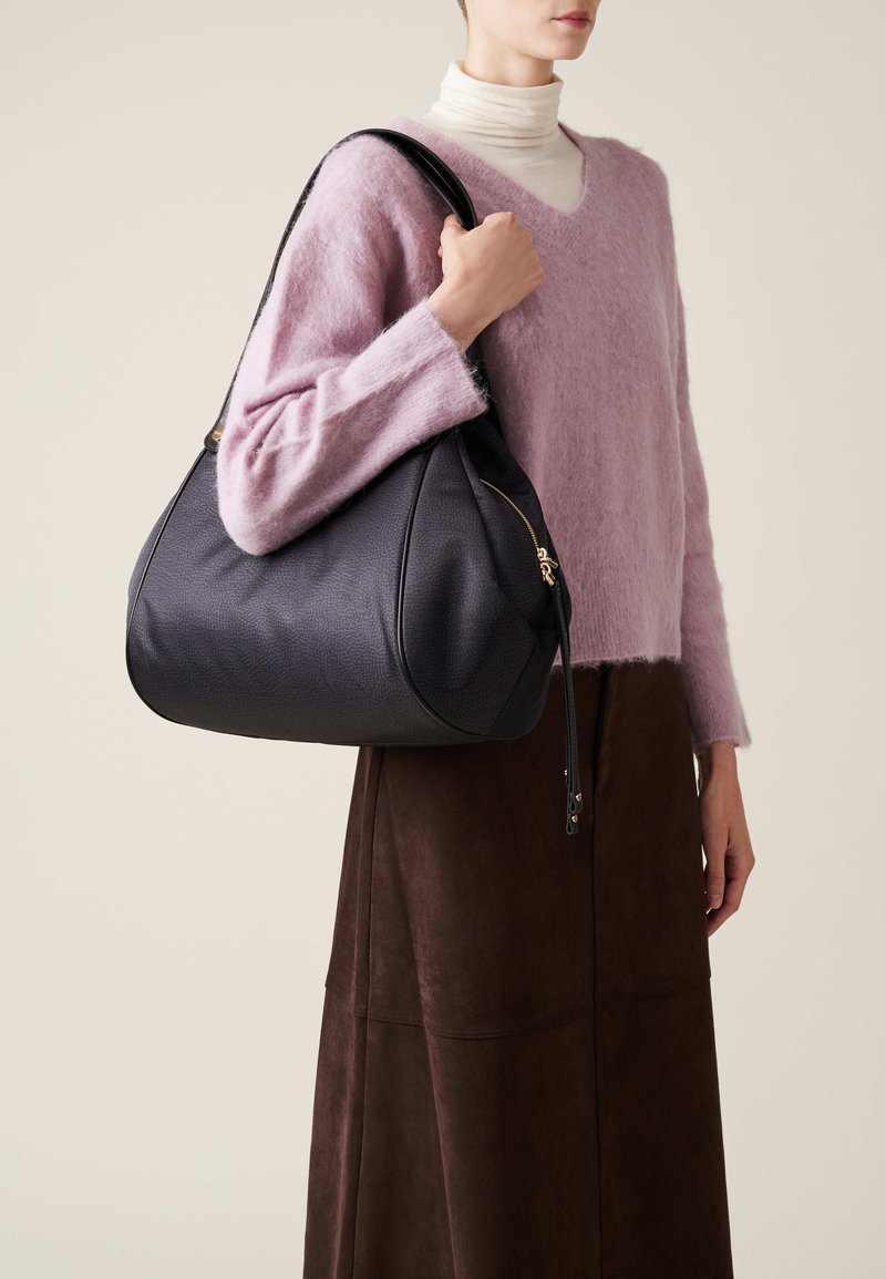 Grande borsa a spalla in pelle blu navy con texture liscia, zip dorate e un design capiente. Indossata sotto un morbido maglione a maniche lunghe rosa.