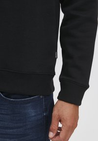 Felpa nera con polsini e orlo a coste, con una piccola etichetta del marchio sul lato. Indossata con jeans in denim blu, che mostrano mano e polso.