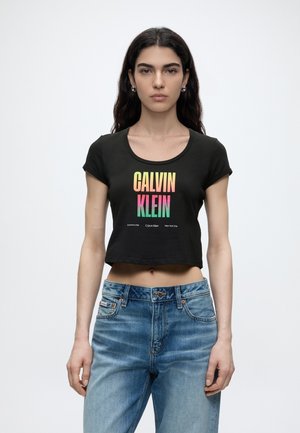 Νεαρή γυναίκα με μακριά σκούρα μαλλιά φοράει μαύρο κοντό μπλουζάκι με πολύχρωμο κείμενο «CALVIN KLEIN» και μπλε τζιν, στέκεται μπροστά σε λευκό φόντο.